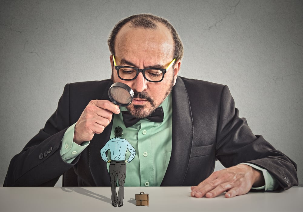 Curious corporate businessman skeptically meeting looking at small employee standing on table through magnifying glass isolated office grey wall background. Human face expression, attitude, perception-1
